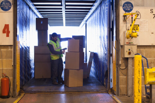 A dock worker loads cardboard boxes onto the back of a truck at a loading bay